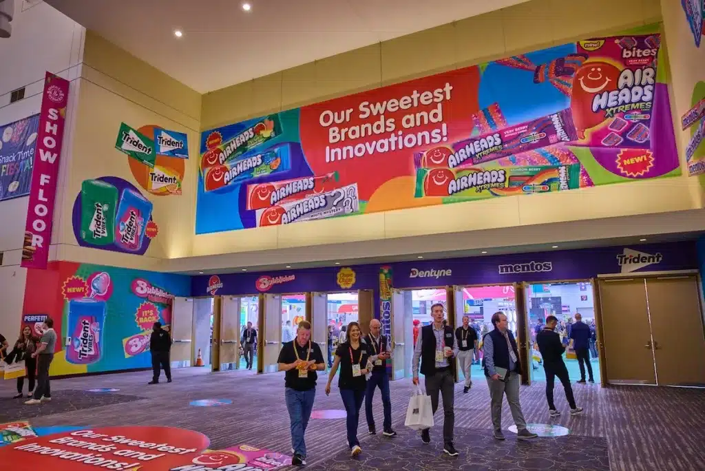 Attendees walking through the Sweets and Snacks Expo entrance hall, with large confectionery brand displays and a Show Floor sign visible.