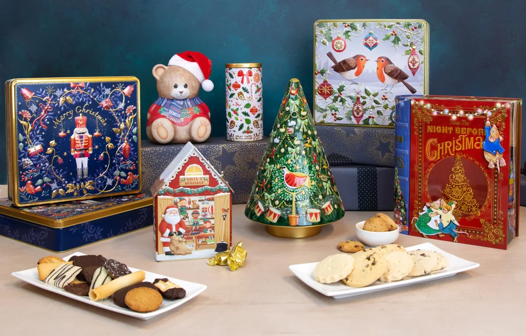 Collection of festive Christmas biscuit tins in various designs including nutcracker, robins, Christmas tree, and Santa's workshop, displayed with biscuits and cookies.