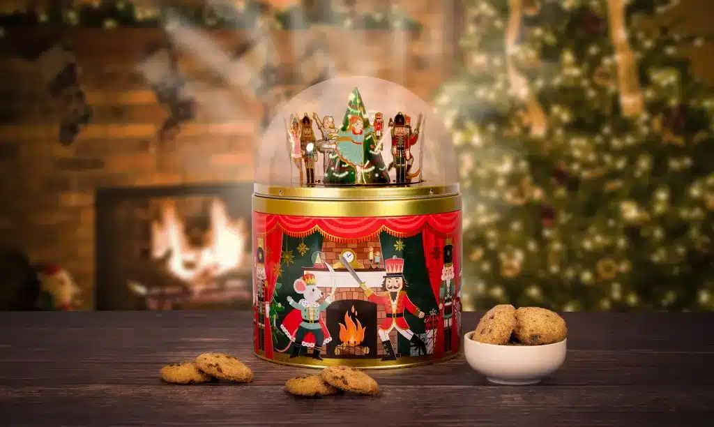Festive Christmas biscuit tin featuring nutcracker characters and snow globe design with cookies displayed beside it.